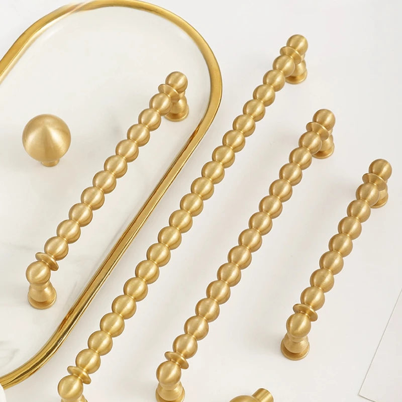 A set of decorative cabinet hardware including long beaded bar handles and round ball knobs in antique bronze, dark bronze, and gold finishes, arranged on a white background with a gray sample board.
