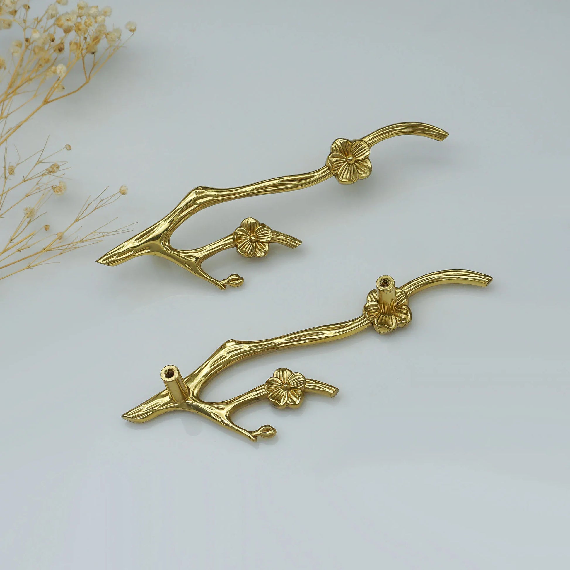 Two gold branch-shaped wall hooks with floral details, displayed on a clean white background with dried baby's breath flowers in the corner.