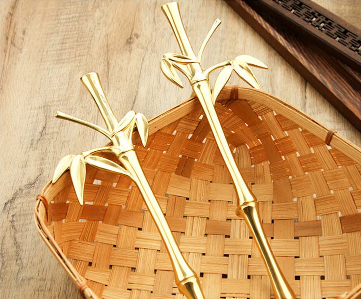 Two gold-toned metal chopsticks with bamboo and leaf designs on the tips, resting inside a woven bamboo basket on a wooden surface.