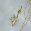 A gold branch-shaped decorative object with sculpted flower details, photographed on a light gray background with dried plant stems along the right side.