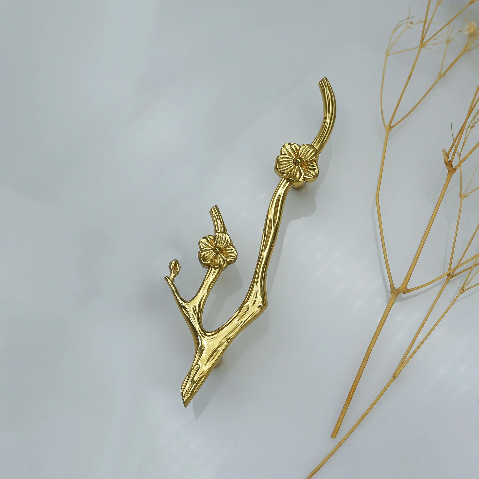 A gold branch-shaped decorative object with sculpted flower details, photographed on a light gray background with dried plant stems along the right side.