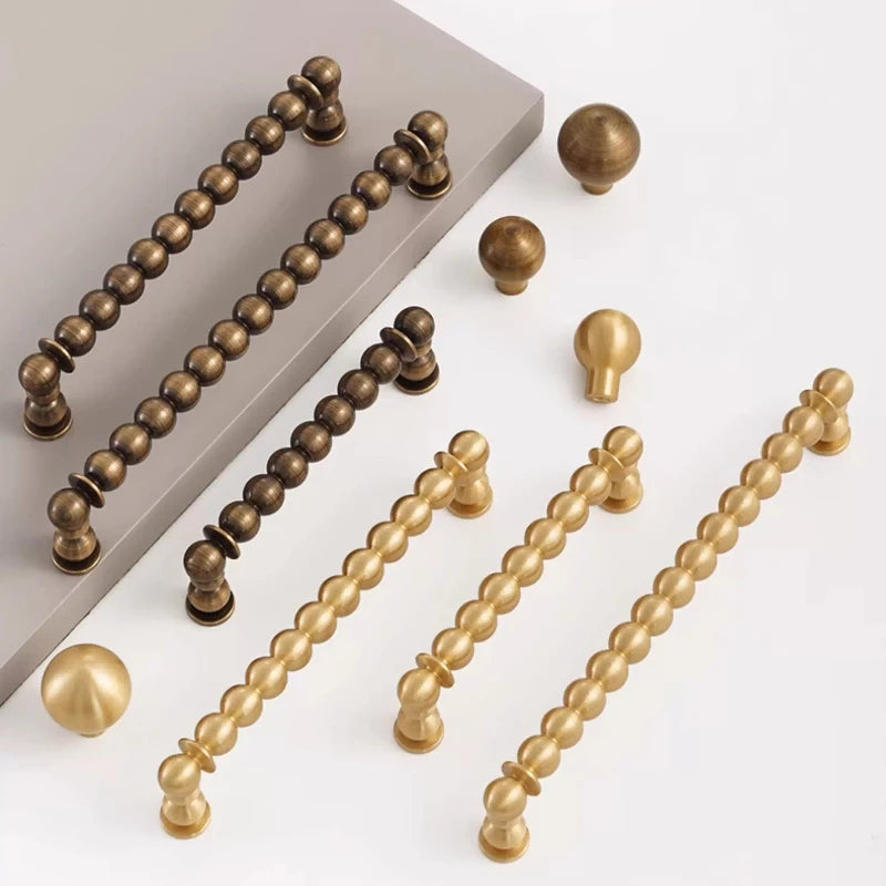 A set of decorative cabinet hardware including long beaded bar handles and round ball knobs in antique bronze, dark bronze, and gold finishes, arranged on a white background with a gray sample board.